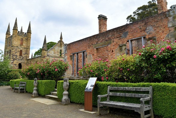 Ruins of a house, built from orange brick. It is mostly just walls, but a chimney can be seen on one side. The front has a doorway, with wide windows either side (typical double-fronted Victorian house).  Fragments of stone decoration are still attached to the wall. 
In front of the house are a row of rose bushes, with pink flowers, and this is fronted by a law, clipped hedge, with two concrete? vases, two benches and a plaque about the house. The "elegance" of the garden contrasts with the roughness of the ruined walls.
Beside the house is the old stone church, now just walls after it was gutted by bushfires. (Presumably this took out the house too.)