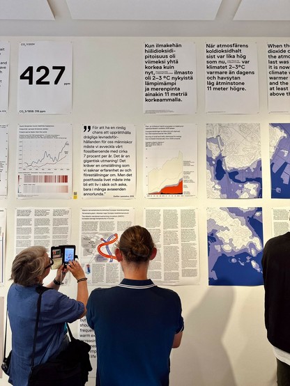 Many posters with text and charts on gallery walls, two onlookers