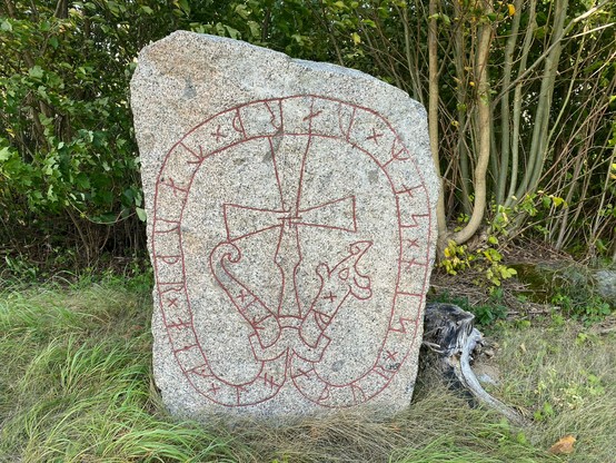 Photo of a runestone with a clear inscription. It has a rune snake going around the edges, connected with a collar at the bottom. Above it is a carved cross.