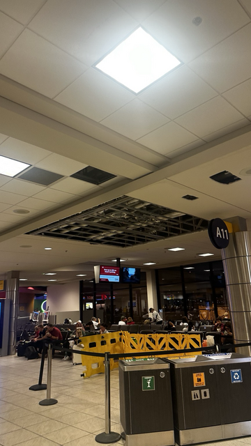 A false ceiling with panels removed and sections cut out, exposing a metal frame and ductwork. A sign for gate A11 is visible.