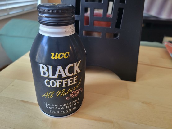 Can of UCC Black Coffee All Natural Unsweetened Coffee Drink, on a birch veneer IKEA table behind a green couch. Partially visible is the side of a matte-black Asian-inspired candle-holder lantern.

Can description:
The can is about 6 inches (15 cm) tall, with a 1-inch (~2.5 cm) neck and 1-inch (~2.5 cm) shoulder. Its label has a black background. Under its black metal cap, the neck is exposed as a short, gray ring showing the best buy date. 

Text on the black background reads: "UCC" in a medium-sized, italicized, yellow with thin gray drop shadow, uppercase, serif block font; "Black Coffee" in a large, off-white with thin yellow drop shadow, uppercase, serif block font; "All Natural" in a medium-sized, yellow, cursive, flowing font in mixed case, rising from left-to-right; "Unsweetened Coffee Drink" in a smaller, white, serif font in mixed case; and "9.74 fluid ounce (288 mL)" in a smaller, white, sans serif block font in mixed case.

Ten photo-realistic coffee beans appear on the right in a loose pile under text "All Natural".