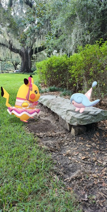 A Pokémon GO AR photograph of Pikachu in the pop star dress standing in front of a Hattarene on a stone bench in a park.