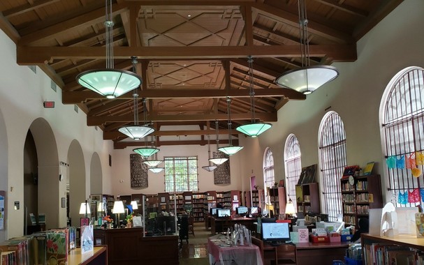 Long view shows wooden high  elevated ceiling with hanging pale blue large simple cone lights and wooden desks, with blue occasional computer monitors and desk lamps, as well as wooden bookcases. Along the sides are very tall arches in the white plaster walls - to the left as doors, to the right, as very tall windows.