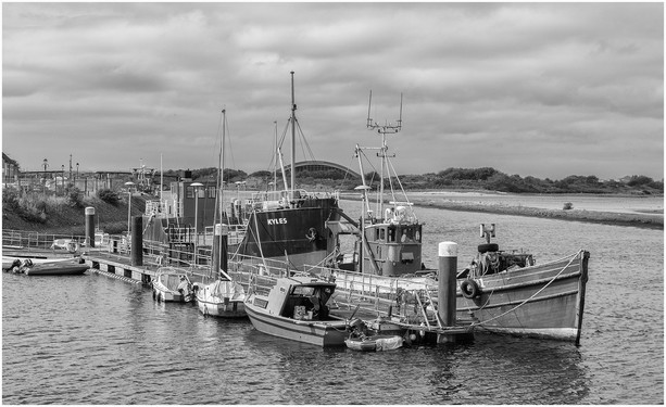 A black and white photo of a dock with several boats, including a larger vessel named "Kyles." The scene features a calm waterway with a cloudy sky and nearby land.