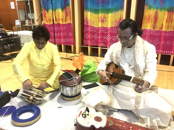 Two seated Indian musicians in formal dress. One tuning a tabla with a small hammer, the other tuning a steel string ukulele.