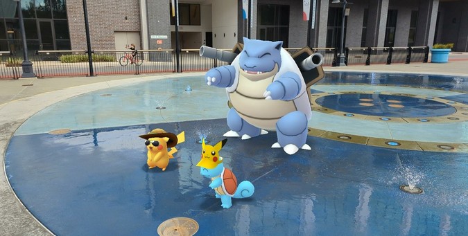 An AR photo taken in Pokémon GO of summer hat Pikachu, Pikachu Visor Squirtle, and Blastoise enjoying a fountain at the local park.