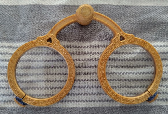 Eine mittelalterliche Nietbrille mit Zierschnitzereien, gefertigt aus Buchenholz, hartgeölt, liegt auf einem grau-weiß gestreiften Küchentuch aus Baumwolle.