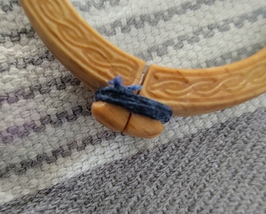 Nahansicht eines Schließblocks der Nietbrille. Dieser besteht aus zwei Holznocken, um die ein dunkelblauer Zwirnfaden gebunden ist. Der Zwirnfaden ist verknotet. Der Knoten befindet sich leicht oberhalb, links am Fadenbündel.

Der Faden ist ebenfalls hartgeölt. Dies lässt sich im Bedarfsfall wieder spurenfrei lösen verstärkt aber auch die Haltbarkeit der Knoten.