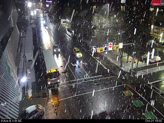 A traffic camera of an urban intersection with snow falling.