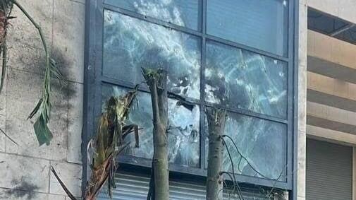 A damaged window with broken glass and palm trees nearby, exhibiting signs of UAV-related impact.