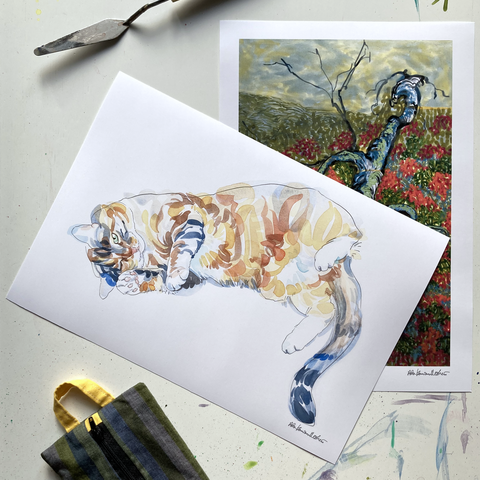 An overhead shot of two small, matte posters laid out on a paint-stained white table top. There is a palette knife poking in from the upper left edge of the image. On the bottom of the image, a green striped cloth pencil case with a yellow loop pokes into frame. The small posters are overlapped, with the bottom poster showing a white-bordered reproduction of an abstracted birch tree in a fall Lapland landscape. The top poster is a reproduction of a cat watercolour painting. The cat is painted loosely in lozenge-shapped blocks of colour. The cat is lounging on its right side, tail curling to the bottom of the paper. The cat has one eye open, as if it is winking at the viewer.