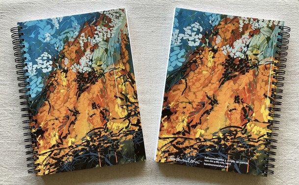 Two spiral-bound notebooks are on an unbleached linen tablecloth. The notebooks covers are printed with the same abstracted midsummer bonfire painting, and the notebooks are showing the front and back covers together. The painting is an eruption of yellow and orange flames against a blue and green background, with the charred silhouettes of the wood and tree branches on the bottom. The other notebook's back cover shows the same bonfire painting with the addition of the artist's signature and painting information on the bottom edge of the notebook.