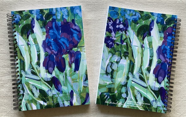 Two spiral-bound notebooks are on an unbleached linen tablecloth. The notebooks covers are printed with the same abstracted flower painting, and the notebooks are showing the front and back covers together. The painting is mostly blue and green, showing the deep blue and purple head of an iris against its leaves and other buds in the background. The other notebook's back cover shows the same blue iris painting with the addition of the artist's signature and painting information on the bottom edge of the notebook.