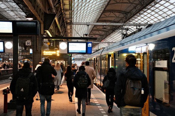 Commuters walk along a bustling train platform in a modern station, with a train on the right. Overhead signs display directions, including "Centrum" and "Hoofddorp." The mood is busy and focused as people move with purpose in the well-lit, spacious setting.