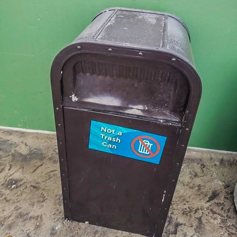 A dark brown container with a sign that reads "Not a Trash Can" accompanied by a prohibition symbol over a trash can icon. The background is a green wall.