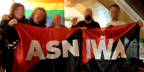 Eine Gruppe Gewerkschafter*innen halten zwei rot-schwarze Fahnen mit der Aufschrift "ASN" und "IWA" vor sich. In Hintergrund hängt an der Wand eine Fortschritts-Prideflagge in Regenbogenfarben.