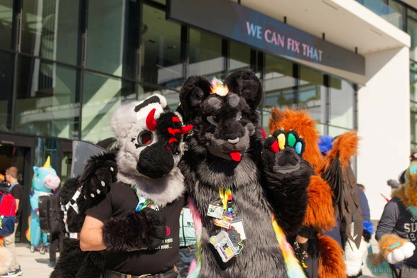 Photo of two fursuiters posing for a photo in front of the CCH entrance. A dragon on the left with light grey fur, black paws and muzzle, and red horns, and a black jaguar with rainbow stripes on the right. Lots of people in the background.