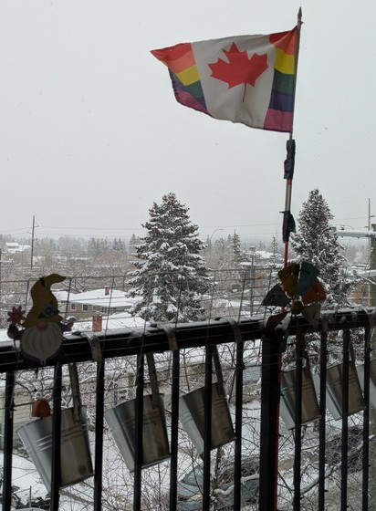 A Pride Canadian flag blows in the wind as the snow starts to fall. 6 silver hanging pots hang off the balcony rail to catch the snow. Fog cuts on the visibility of the horizon with snow covered rooftops and trees in the background.