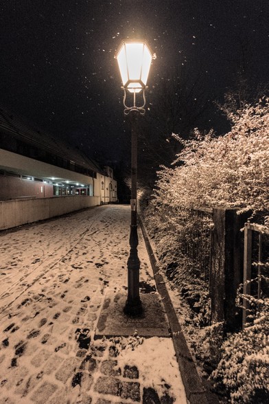 Weitwinkelfotografie im Hochformat. Das Bild zeigt eine leuchtende Straßenlaterne in der späten Abenddämmerung, die vor einem dunkelblauen Himmel in einem Schneegestöber gelblich leuchtet. Die Schneeflocken schweben um die Laterne herum in deR Luft. Auf dem Boden liegt eine dünne weiße Schneeschicht, die in das leicht gelbliche Licht der Laterne getaucht ist. Rechts am Weg nach hinten entlang verläuft ein Metallzaun, dahinter sind schneebedeckte Büsche. Weiter hinten ist links ein Gebäude zu erkennen.