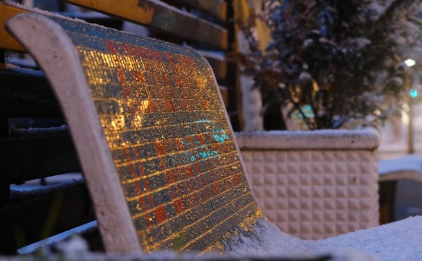 Leicht seitliche Fotografie einer Steinbank in der Abenddämmerung. Die Lehne der Bank ist mit grünen und roten Mosaiksteinen besetzt, aber deren Farbe ist über die Reflektionen von gelben und Ampel-grünen Straßenlichtern nicht so leicht zu erkennen. Die Sitzfläche ist mit Schnee bedeckt. Hinter der Bank ist ein steinerner Blumenkübel mit einem schneebedeckten Strauch zu sehen.