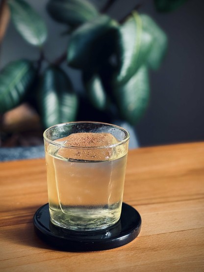 light yellow crystal clear cocktail with big rock of clear ice and nutmeg garnish sitting on a black ceramic coaster on a beechwood table with plants in the background