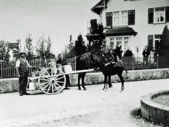 Schwarz-weißes Foto von einem Milchmann, der mit einem Gespann aus einem Pferd und einem Fohlen vor einem Haus steht. Der Milchmann steht neben einem einspännigen Karren, der mit Milchkannen beladen ist. Ein weiterer Mann steht in der Nähe des Karrens. Im Hintergrund ist ein Haus mit Gartenzaun zu sehen. Das Bild wirkt historisch.