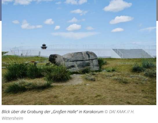 Ansicht über die Grabung der "Großen Halle" in Karakorum. Im Vordergrund eine Steinskulptur in Steppenlandschaft, dahinter eine neu errichtete Plattform mit Treppenstufen.