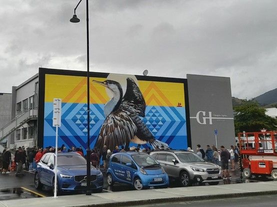 The wall of a previously grey lawyer building is painted with a large mural featuring a bird. The top third of the background is the sky layer, yellow and orange from the sun (which is behind the birds head). The bottom two thirds are ocean blue, with the bottom one darker than the middle. Throughout them are patterns using triangles wrapped in two outlines of diamond shapes. 
In front of the mural is a group of people attending the opening ceremony, and some cars are parked in the carpark in front.