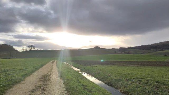 Hegaulandschaft mit Weg, Wiesen und Bergen. Die Sonne bricht durch die Wolken und es entstehen sichtbare Lichtstrahlen.