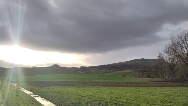 Hegaulandschaft mit Bachlauf, Wiesen und Bergen. Die Sonne bricht durch die Wolken und es entstehen sichtbare Lichtstrahlen.