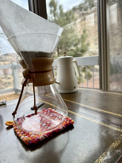 An empty Chemex with a square filter full of grounds in the top. It’s resting on a crocheted pad on a leather-topped desk. Behind it is a white, goose necked kettle. Behind that is a window, through which can be seen a deck railing, the top of a pine tree, and sandstone cliffs in the distance.