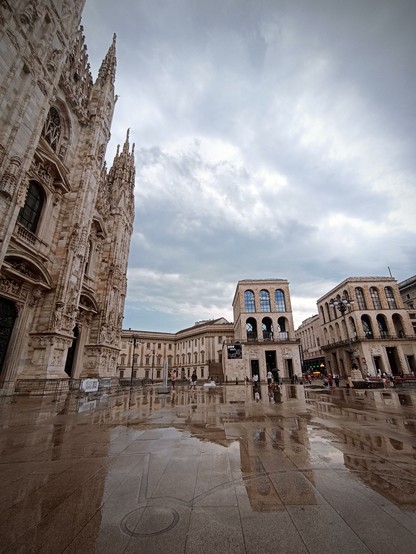 Fotografía de la plaza del Duomo de Milán. A la izquierda la fachada del Duomo, en frente el museo y otros edificios. El cielo está nublado y el suelo mojado por la lluvia.