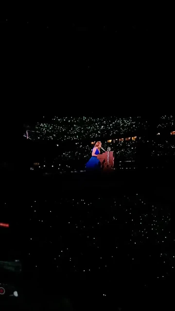 Captura de un vídeo de Taylor Swift sentada en el piano de las canciones sorpresa con la guitarra delante. Ella lleva un vestido azul y el estadio está lleno de luces verdes.