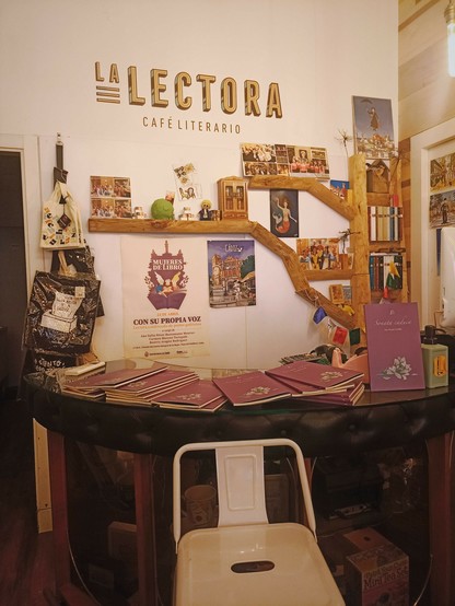 Fotografía de una mesa negra semicircular con varios ejemplares del libro "Sonata caduca" dispuestos a lo largo de la misma. Estos libros son morados con una ilustración de un nardo formado por partituras. Delante de la mesa hay un taburete blanco. Detrás una pared blanca con varios carteles y estanterías en la que destaca el rótulo amarillo "LA LECTORA. CAFÉ LITERARIO".