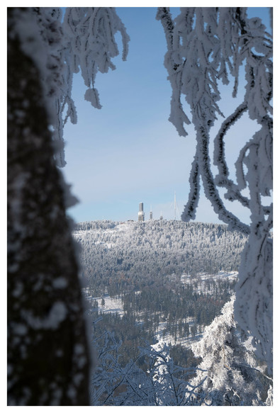 The photo shows a snow-covered and wooded mountain in the Taunus. There are three towers and a transmission mast on the mountain. The photo was taken through a tree, so that ice-covered branches can be seen in the foreground.