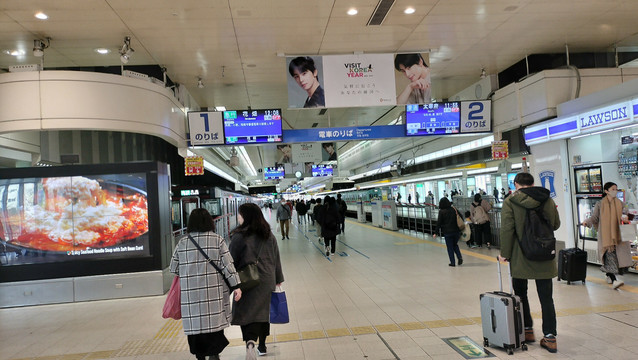 A photo of Nishitetsu Fukuoka Station. A walkway recedes into the distance. On either side of the walkway are the train platforms; the left side platform has a train waiting. People are walking up and down the walkway. An electronic billboard advertising some hot noodles is on the left side in front of where the train platform begins, and a Lawson convenience store is in front of the right side platform.