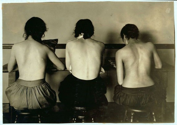 The image depicts three individuals sitting at a table with their backs to the camera. They appear to be in an indoor setting, possibly a classroom or office environment from a bygone era given the style of clothing and furniture.

All three subjects are wearing what looks like dark-colored shorts over skirts that reach down to just above their knees. Their hair is pulled back away from their faces, suggesting they have taken off any headwear for comfort during the activity at hand.

The individuals seem engaged in writing or drawing on a sheet placed on the table before them. The lighting of the photo casts soft shadows behind and beneath each person's legs as well as under the furniture, indicating an interior space with artificial light sources such as lamps or overhead lights.

The monochrome tone of this image suggests it could be historical in nature; possibly from early 20th-century America given its style. The individuals' postures appear relaxed yet attentive to their task at hand.