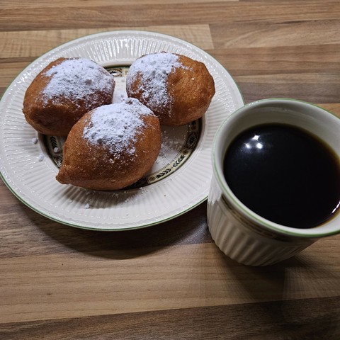 Foto van drie, grote, oliebollen op een ontbijtbord, bestrooid met poedersuiker. Ernaast een grote mok koffie.