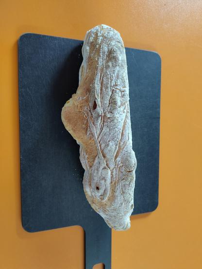 Ein kleines Baguette liegt auf einem schwarzen Schneidbrett, das liegt auf einer orangenen Arbeitsfläche.