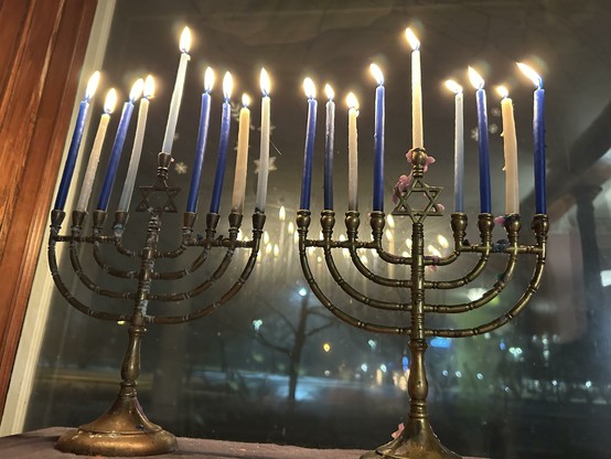 Two traditional brass chanukiyahs fully lit with a mixture of white and blue candles. The window is dark beyond.