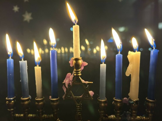 A brass chanukiyah in front of a dark window, with blue and white candles more than halfway burnt down. The flames reflect in the window. The chanukiyah itself is in shadow.