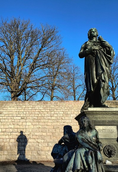 Eine sonnige Szene in einem Park. Vorne rechts ist ein massives Bronzedenkmal mit einem in ein Gewand gehüllten Friedrich Schiller. Er steht auf einem quadratischen Sockel. Unterhalb sitzen um ihn herum vier ebenfalls massive, übergroße Personen in Gewändern.  Auf dem Sockel steht "Schiller". Etwas weiter hinten befindet sich eine hohe Mauer aus hellen Steinen. Darauf ist unten links der Schattenriss der Schillerskulptur zu sehen. Hinter der Mauer stehen einige kahle Winterbäume vor einem knallblauen wolkenlosen Winterhimmel.

A sunny scene in a park. In front on the right is a massive bronze monument with a robed Friedrich Schiller. He stands on a square pedestal. Below him, four equally massive, oversized figures in robes are seated around him.  "Schiller" is written on the plinth. A little further back is a high wall of light-coloured stones. The silhouette of the Schiller sculpture can be seen at the bottom left. Behind the wall are some bare winter trees against a bright blue, cloudless winter sky.