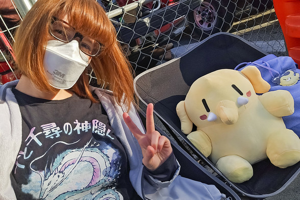 Photo of Lety and the Creature Mastodon stuffed toy in their seats and waiting for the Rose Parade. From top to bottom, she is seen wearing a light blue winter jacket and a graphic tee featuring Haku from Spirited Away and the text  “千と千尋の神隠し”. Lety's in the foreground, only the upper half of her body visible, sitting on a small black folding chair and smiling at the camera as she makes a peace sign with her left hand. Beside her is the Creature sitting on a matching chair next to its tote bag, and further behind their backs is a wire metal fence.