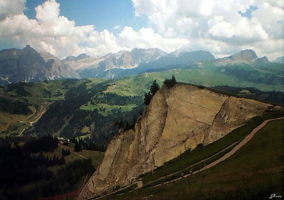 Landschaftsfoto in Farbe, aufgenommen in den Dolomiten über Corvara. Im Vordergrund rechts ein beiger Fels, zu dem ein Fußweg hinaufführt. Weiter entfernt mittelhohe grüne Berge, teilweise mit Wald, am Horizont Hochgebirge und darüber weiße Wolken am blauen Himmel. Diana Mini auf Kodak Gold.