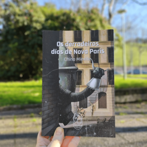 Uma mão branca sostem o livro 'Os derradeiros dís de Nova París", de China Miévielle. De fundo, um parque com relva e árvores verdes.