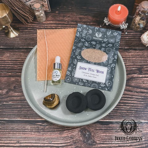 The contents of our exclusive "Snow Full Moon Ritual Kit for the February 12, 2025, Full Moon in Leo" product are arranged on a small tray. They include a sheet of orange beeswax and a wick; a pouch of Snow Full Moon herbs, including cinnamon, orange peel, and St. John's wort; a dropper vial of our exclusive Full Moon Spell Oil; a tumbled tiger's eye gemstone; and two charcoal discs. Presented by Inked Goddess Creations.