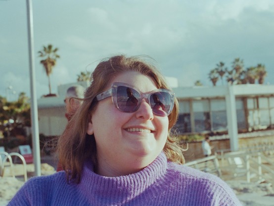 my girlfriend looking to the side and smiling. she is wearing a purple sweater and sunglasses, and behind her are some palm trees and clouds. you can see me in my red coat in the reflection in her glasses.
the photo has old-school-photos muted colours resulting from the combination of the film and lighting.