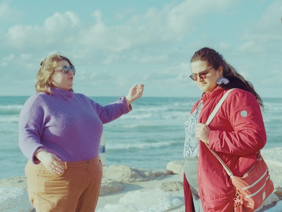 my girlfriend and i taking while standing on the beach. she is wearing a purple sweater and sunglasses, i a red coat, transition glasses and trans-coloured earrings, and a large red purse. behind us are waves and clouds.
the photo has old-school-photos muted colours resulting from the combination of the film and lighting.