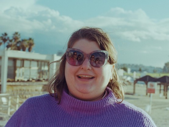 my girlfriend looking at the camera and smiling. she is wearing a purple sweater and sunglasses, and behind her are some palm trees and clouds.
in the reflection in her glasses you can see me in my red coat and Utna taking the picture.
the photo has old-school-photos muted colours resulting from the combination of the film and lighting.
