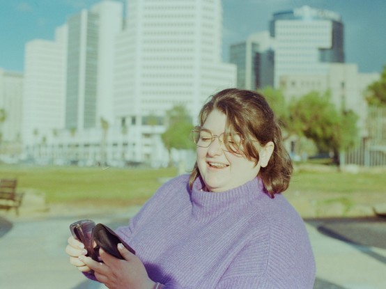 my girlfriend being blinded by the sun as she is switching over to sunglasses. she is wearing a purple sweater and glasses, and behind her are some beachfront buildings.
the photo has old-school-photos muted colours resulting from the combination of the film and lighting.